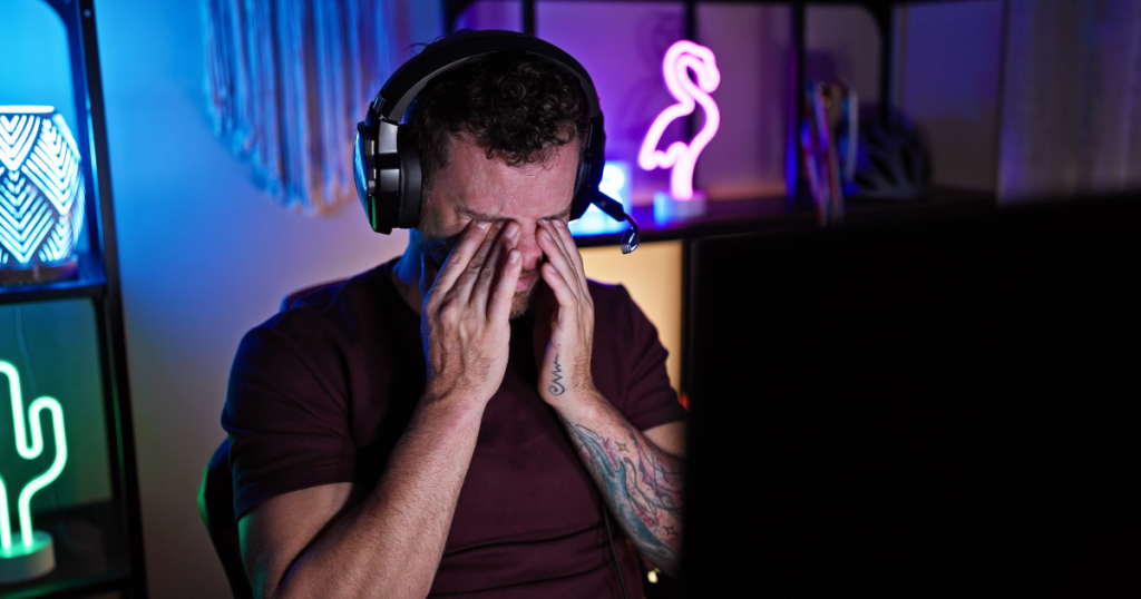 Stressed gamer rubbing forehead with a lit screen in the background. Mid-season burnout starting to hit